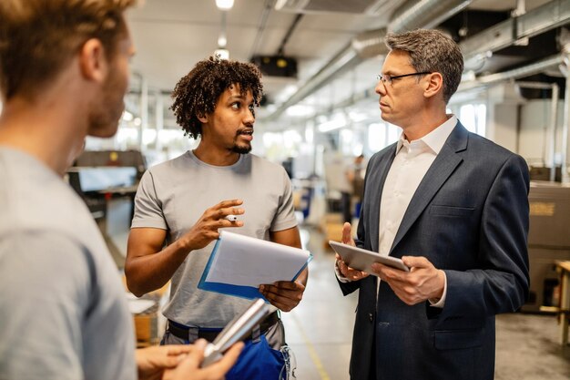 Ingeniero adulto medio que usa un panel táctil y se comunica con un trabajador afroamericano sobre informes de productos en edificios industriales