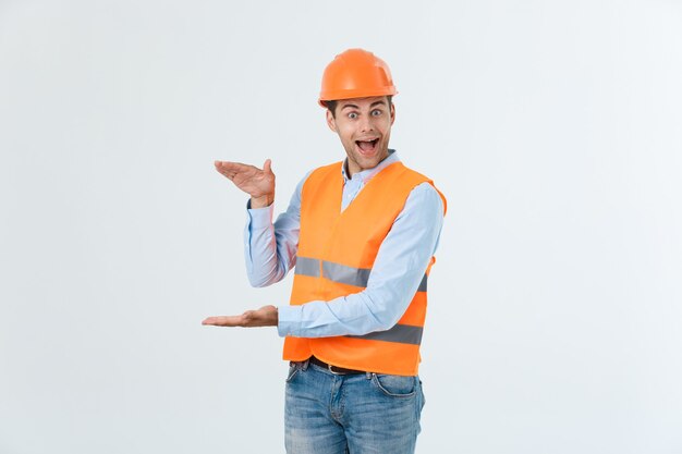 Ingeniero de barba feliz sosteniendo la mano a un lado y explicando algo, chico vestido con camisa caro y jeans con chaleco amarillo y casco naranja, aislado sobre fondo blanco.