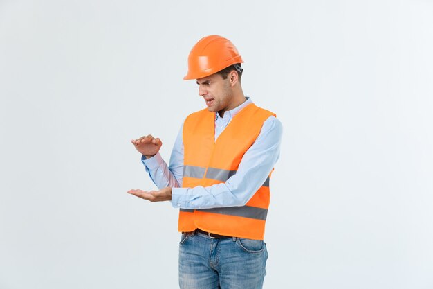 Ingeniero de barba feliz sosteniendo la mano a un lado y explicando algo, chico vestido con camisa caro y jeans con chaleco amarillo y casco naranja, aislado sobre fondo blanco.
