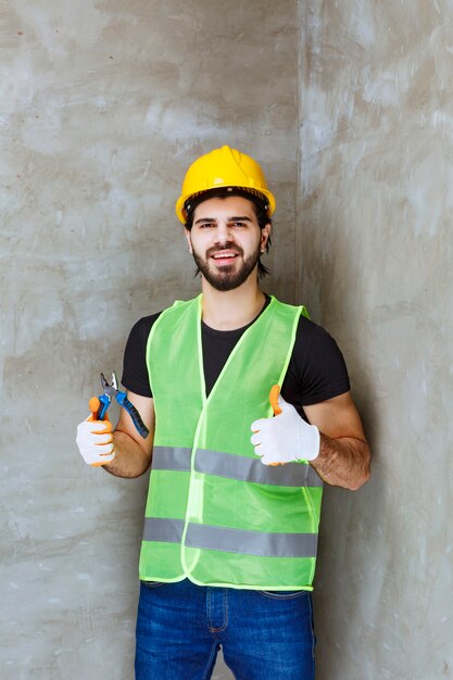 Ingeniero en casco amarillo y guantes industriales sosteniendo alicates azules y mostrando el pulgar hacia arriba signo