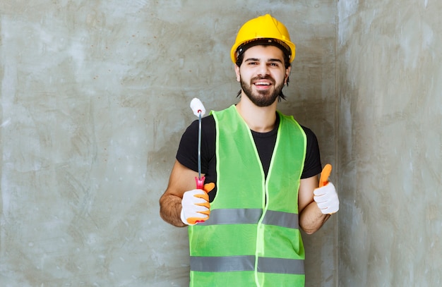 Ingeniero con casco amarillo y guantes industriales sosteniendo sosteniendo un rodillo de pintura y disfrutando del producto