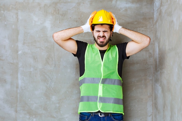 Ingeniero con casco amarillo y guantes industriales tratando de quitarse el casco ya que tiene dolor de cabeza