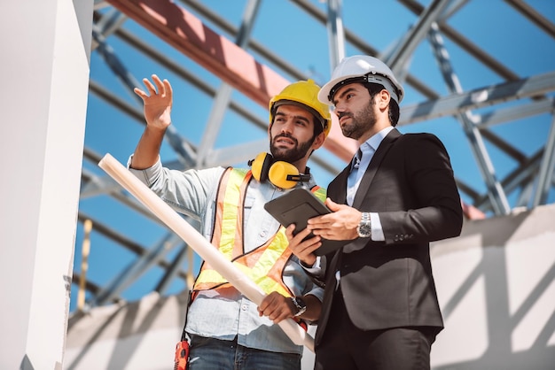 Ingeniero civil y gerente de trabajadores de la construcción con tableta digital y planos hablando y planeando sobre el sitio de construcción Concepto de trabajo en equipo de cooperación