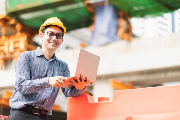 Ingeniero civil profesional masculino asiático inteligente control de construcción del sitio de la computadora portátil con infraestructura y antecedentes de construcción