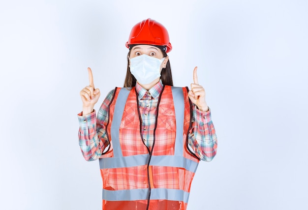 Ingeniero de construcción femenino en casco rojo y máscara de seguridad apuntando hacia arriba.