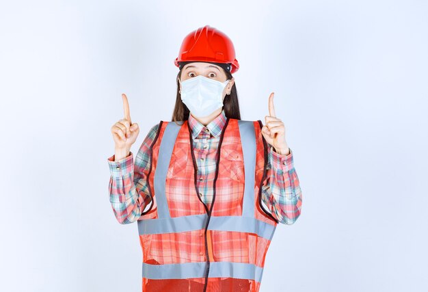 Ingeniero de construcción femenino en casco rojo y máscara de seguridad apuntando hacia arriba.