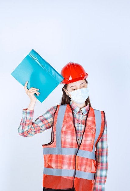 Foto gratuita ingeniero de construcción femenino en máscara de seguridad y casco rojo sosteniendo una carpeta azul y parece confundido y pensativo