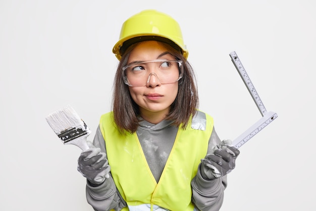 Ingeniero de construcción de mujer asiática pensativa en uniforme sostiene cinta métrica para medir el diseño y pincel listo para trabajar en la construcción de algo contra la pared blanca. Trabajador industrial