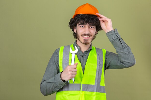 Ingeniero guapo joven barbudo que sostiene la llave tocando el casco con la mano con una sonrisa en la cara sobre la pared verde aislada