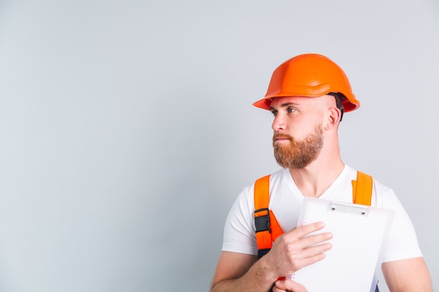 Foto gratuita ingeniero de hombre guapo en la construcción de casco protector en la pared gris tiene portapapeles blanco