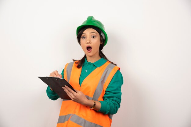 Ingeniero industrial mujer sorprendido en uniforme con portapapeles sobre fondo blanco.