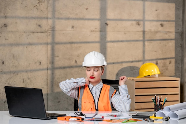 Ingeniero inteligente joven linda chica rubia trabajador civil en casco y chaleco cansado sosteniendo el cuello