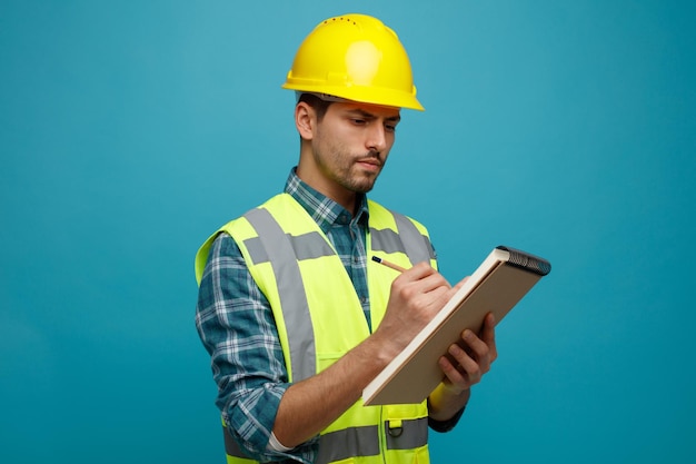 Ingeniero joven concentrado con casco de seguridad y uniforme de pie en la vista de perfil sosteniendo lápiz y bloc de notas tomando notas de trabajo aisladas en fondo azul