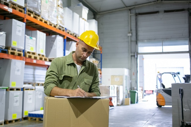 Ingeniero logístico masculino serio en casco y posición general en la pila de cajas y completando documentos. Copie el espacio. Concepto de cuello azul o trabajo