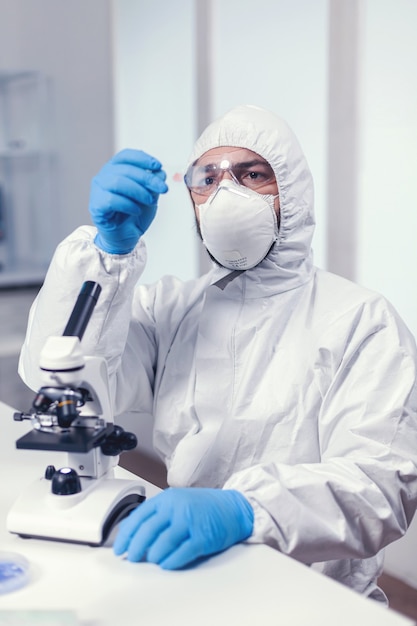 Foto gratuita ingeniero médico mirando atento a la muestra en portaobjetos de vidrio sentado en su lugar de trabajo vestido con traje de ppe