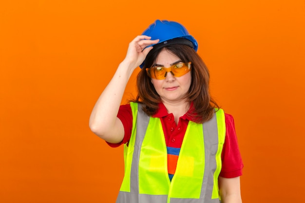 Ingeniero mujer con gafas de chaleco de construcción y casco de seguridad tocando el casco con la mano como gesto de saludo sobre pared naranja aislada