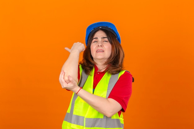 Ingeniero mujer vistiendo chaleco de construcción y casco de seguridad mirando mal tocar el codo con dolor de pie sobre la pared naranja aislada