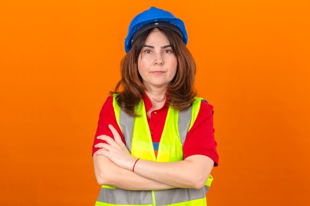 Ingeniero mujer vistiendo chaleco de construcción y casco de seguridad de pie con los brazos cruzados con cara seria sobre pared naranja aislada