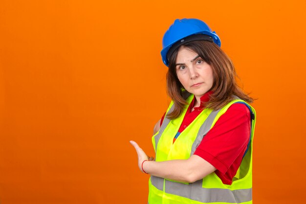 Ingeniero mujer vistiendo chaleco de construcción y casco de seguridad que indica algo en el espacio en blanco de la copia detrás de ella sobre la pared naranja aislada