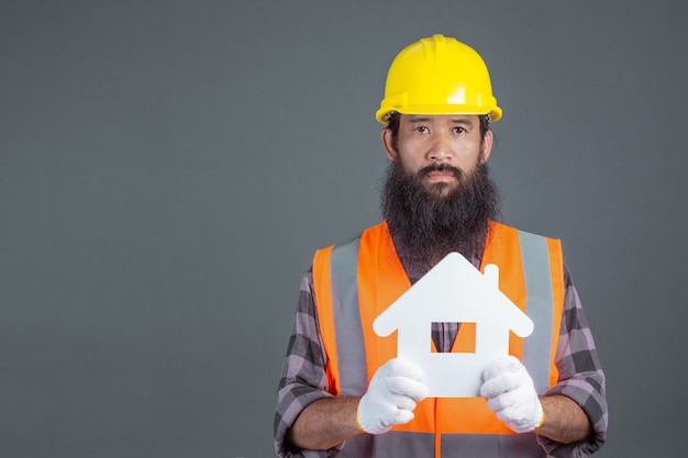 Un ingeniero de sexo masculino que lleva un casco de seguridad amarillo que lleva a cabo un símbolo de la casa blanca en un gris.