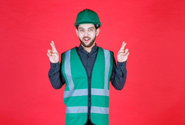 Ingeniero en uniforme verde y casco con signo de cruz de dedo.