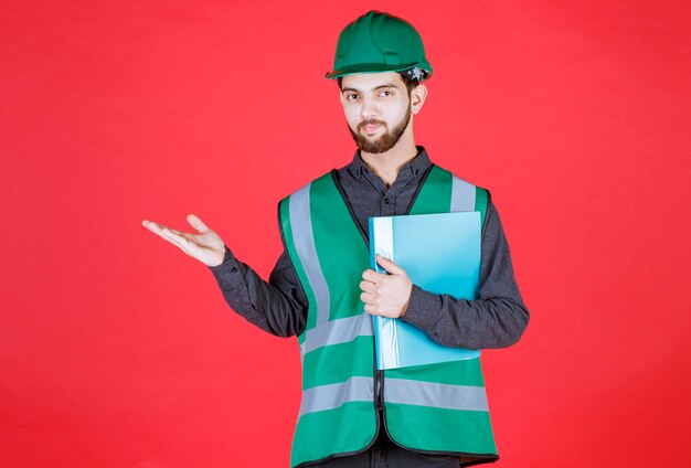 Ingeniero en uniforme verde y casco sosteniendo una carpeta azul y apuntando algo a la izquierda.