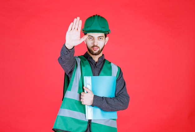 Ingeniero con uniforme verde y casco sosteniendo una carpeta azul y deteniendo a alguien.