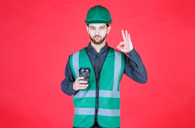 Ingeniero en uniforme verde y casco sosteniendo una taza de café desechable negra y disfrutando del sabor.