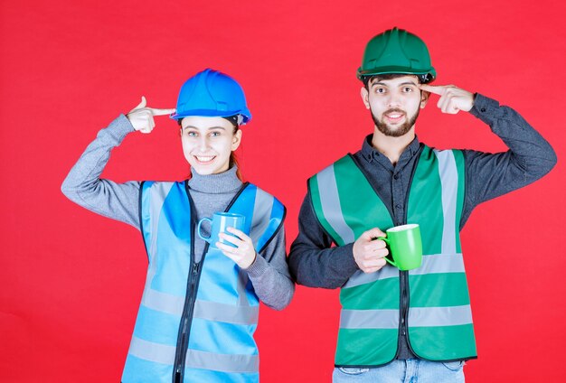 Ingenieros masculinos y femeninos con casco sosteniendo tazas azules y verdes y pensando en nuevas ideas.