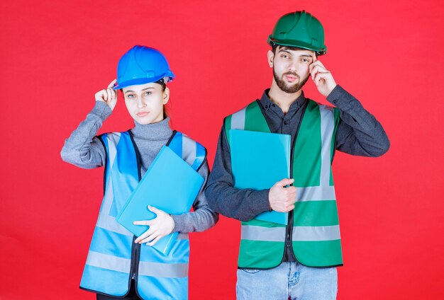 Ingenieros masculinos y femeninos con cascos sosteniendo carpetas azules y parecen confundidos y pensativos.