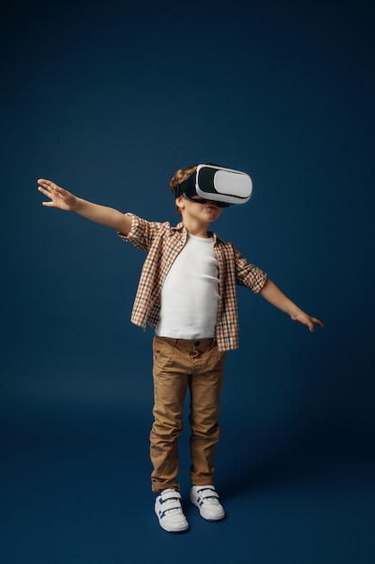 Ingrávido en el cielo. Niño o niño en jeans y camisa con gafas de casco de realidad virtual aisladas sobre fondo azul de estudio. Concepto de tecnología de punta, videojuegos, innovación.