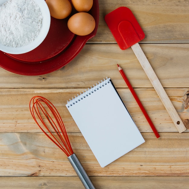 Ingredientes de los pasteles en la placa con los utensilios y la libreta espiral en superficie de madera