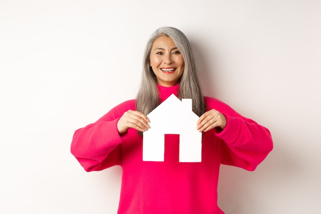 Inmobiliaria hermosa abuela asiática mostrando casa de papel y sonriendo feliz de pie en sudor rosa ...