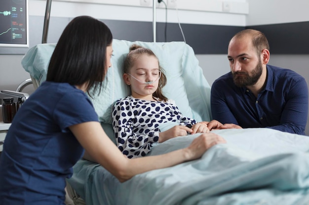 Inquietos e inquietos padres cuidadosos que conversan sobre la evolución de la enfermedad de las niñas pequeñas mientras se encuentran en la sala de recuperación de pacientes del hospital pediátrico. Jóvenes nerviosos preocupados hablando sobre el tratamiento de la salud