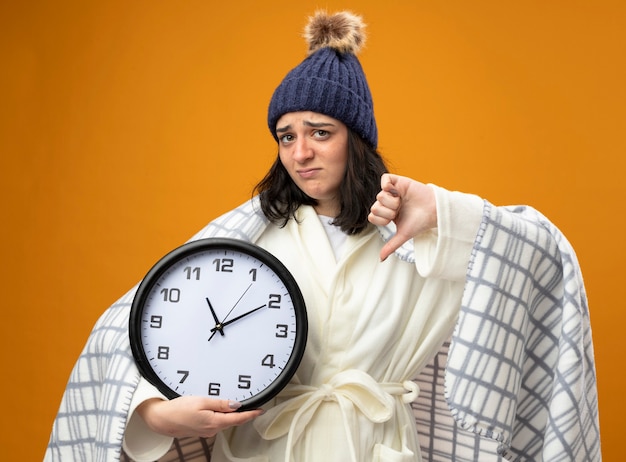 Insatisfecha joven enferma vistiendo bata sombrero de invierno envuelto en cuadros sosteniendo el reloj mirando al frente mostrando el pulgar hacia abajo aislado en la pared naranja