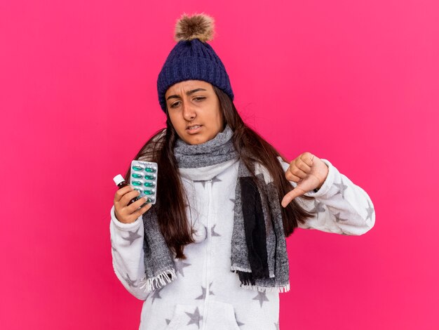 Insatisfecha joven enferma vistiendo gorro de invierno con bufanda sosteniendo la medicina en botella de vidrio mostrando el pulgar hacia abajo aislado sobre fondo rosa