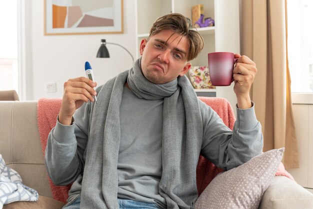 Insatisfecho joven enfermo con bufanda alrededor del cuello sosteniendo el termómetro y la taza sentado en el sofá en la sala de estar