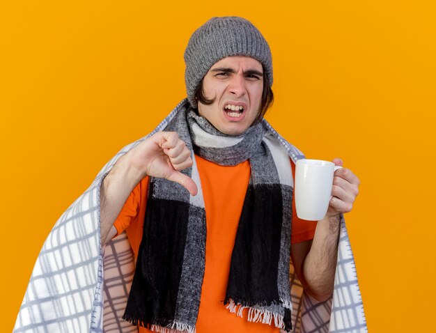 Insatisfecho joven enfermo con sombrero de invierno con bufanda envuelta en cuadros sosteniendo una taza de té mostrando el pulgar hacia abajo aislado sobre fondo naranja