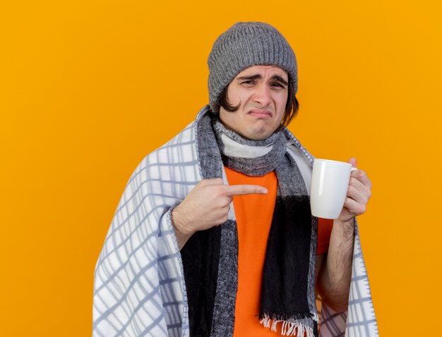 Insatisfecho joven enfermo con sombrero de invierno con bufanda envuelta en plaid sosteniendo y apunta a una taza de té aislado sobre fondo naranja