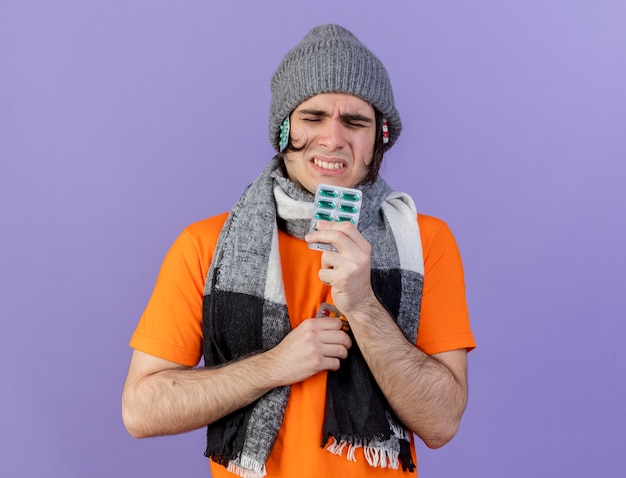 Insatisfecho joven enfermo con sombrero de invierno con bufanda poniendo píldoras debajo del sombrero y sosteniendo píldoras aisladas en púrpura