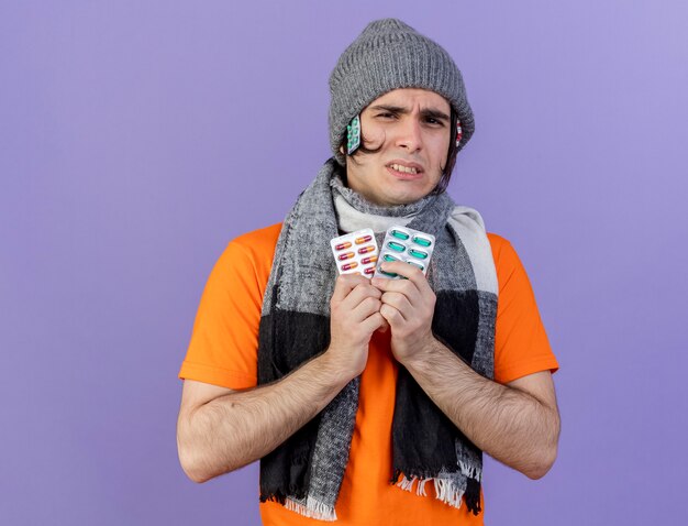 Insatisfecho joven enfermo con sombrero de invierno con bufanda poniendo píldoras debajo del sombrero y sosteniendo píldoras aisladas en púrpura