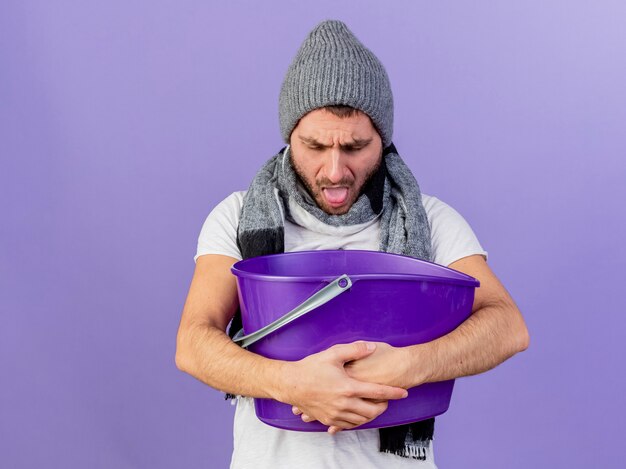 Insatisfecho joven enfermo con sombrero de invierno con bufanda sosteniendo un cubo de plástico y vomitando en él aislado sobre fondo púrpura