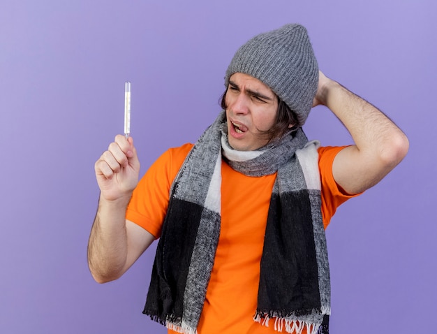 Insatisfecho joven enfermo con sombrero de invierno con bufanda sosteniendo y mirando el termómetro poniendo la mano en la cabeza aislada sobre fondo púrpura