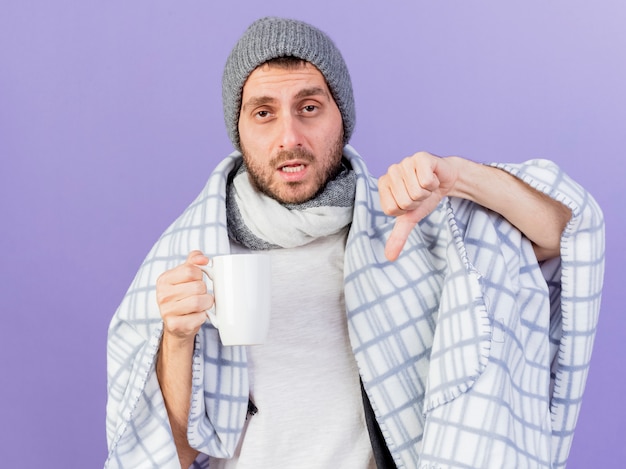 Foto gratuita insatisfecho joven enfermo con sombrero de invierno con bufanda sosteniendo una taza de té mostrando el pulgar hacia abajo aislado sobre fondo púrpura