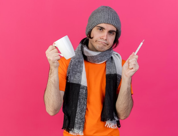 Insatisfecho joven enfermo con sombrero de invierno con bufanda sosteniendo una taza de té con termómetro aislado en rosa