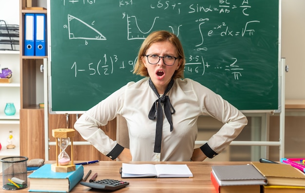 Insatisfecho joven profesora con gafas se sienta a la mesa con útiles escolares poniendo las manos en la cadera en el aula