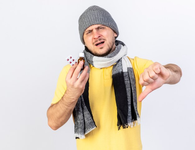 Insatisfecho joven rubio enfermo con sombrero de invierno y bufanda con los pulgares hacia abajo y sostiene la medicina en una botella de vidrio y un paquete de píldoras médicas aisladas en la pared blanca