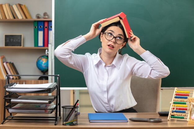 Insatisfecho mirando hacia arriba la joven maestra con gafas cubrió la cabeza con el libro sentado a la mesa con herramientas escolares en el aula