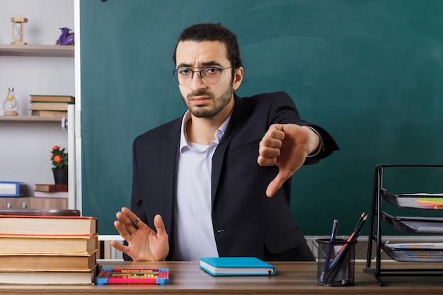 Insatisfecho mostrando el pulgar hacia abajo profesor con gafas sentado a la mesa con herramientas escolares en el aula