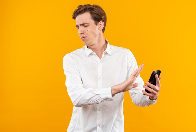 Insatisfecho con los ojos cerrados joven guapo con camisa blanca sosteniendo el teléfono aislado en la pared naranja